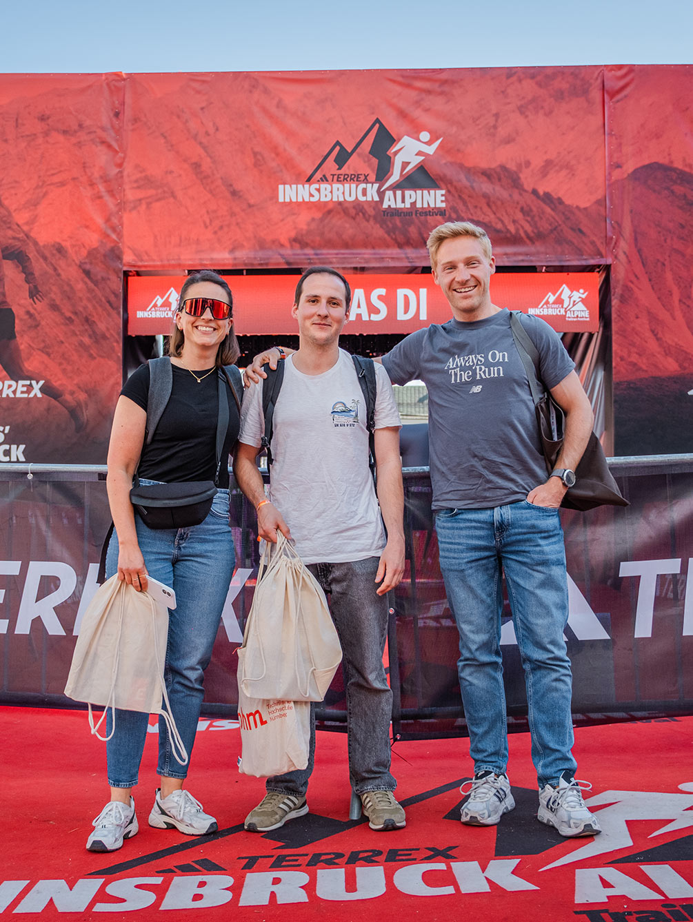 Drei Freunde stehen lächelnd mit ihren Starterbags vor dem offiziellen roten IATF-Zielbogen in Innsbruck.