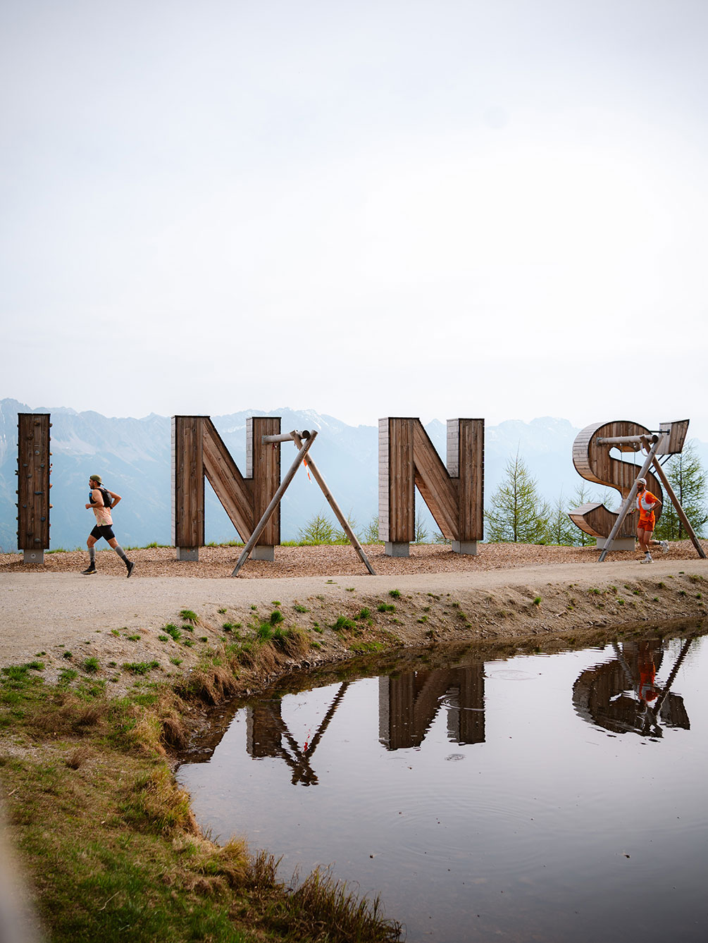 Zwei Trailrunner laufen an den großen hölzernen INNS-Buchstaben an einem Bergsee vorbei, mit den Alpen im Hintergrund während des Innsbruck Alpine Trailrun Festivals.
