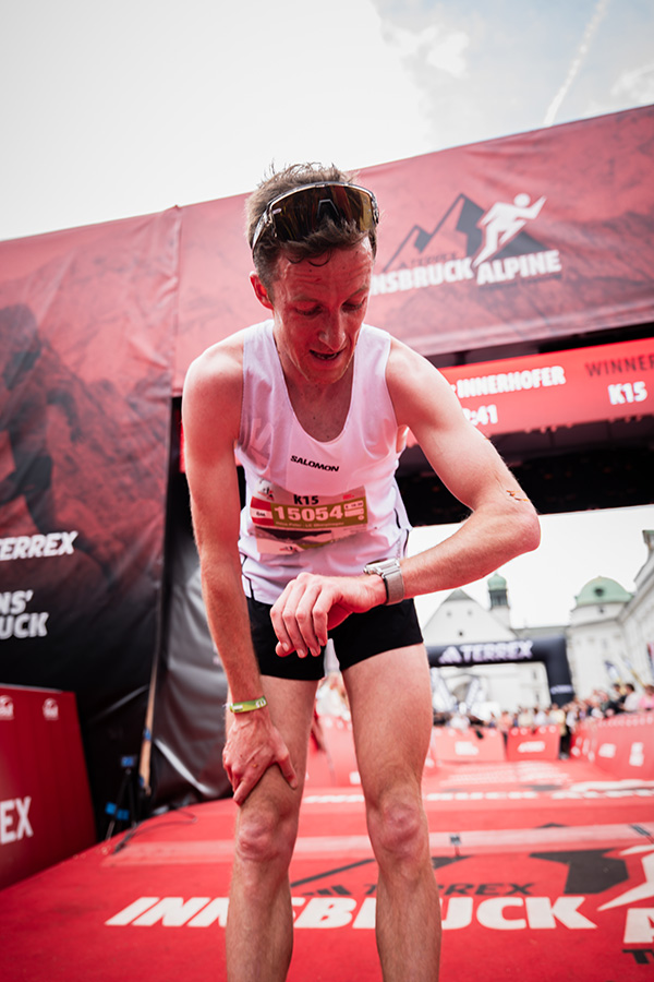 Ein erschöpfter Trailrunner blickt direkt nach dem Zieleinlauf des IATF K15 auf seine Sportuhr; im Hintergrund ist der Zielbogen und das Panorama von Innsbruck zu sehen.
