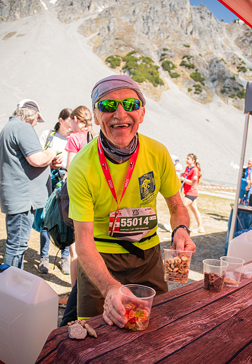 Ein strahlender älterer Trailrunner greift an einer Verpflegungsstation nach Bechern mit Gummibärchen und Brezeln vor einer alpinen Bergkulisse.