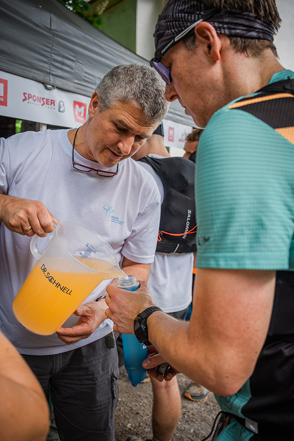 Ein Volunteer an einer Versorgungsstation befüllt die Trinkflasche eines Trailrunners beim Innsbruck Alpine Trailrun Festival mit einem Sportgetränk.