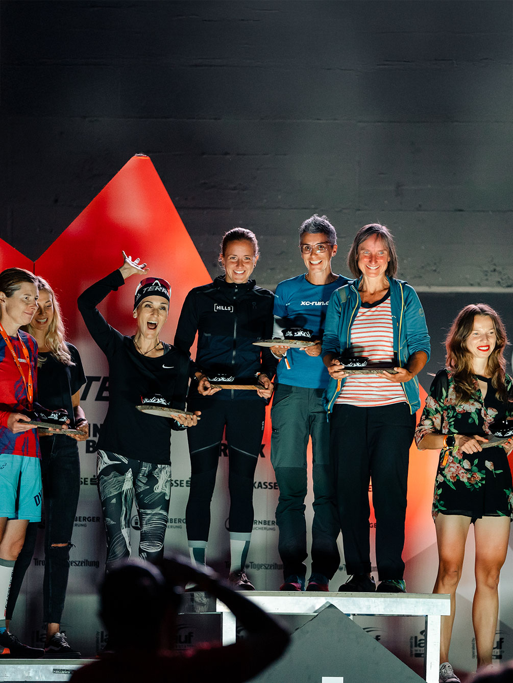 Siegerehrung der Damen beim Innsbruck Alpine Trailrun Festival, bei der sieben strahlende Läuferinnen ihre Trophäen auf dem Podium präsentieren.
