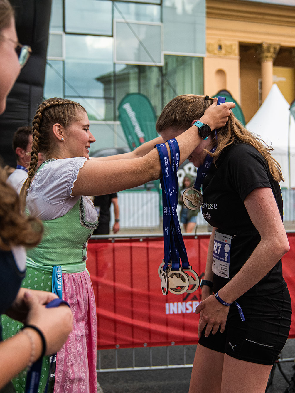 Eine junge Frau in traditionellem Tiroler Dirndl überreicht einer glücklichen Trailrunnerin im Zielbereich des Innsbruck Alpine Trailrun Festivals eine Finisher-Medaille.