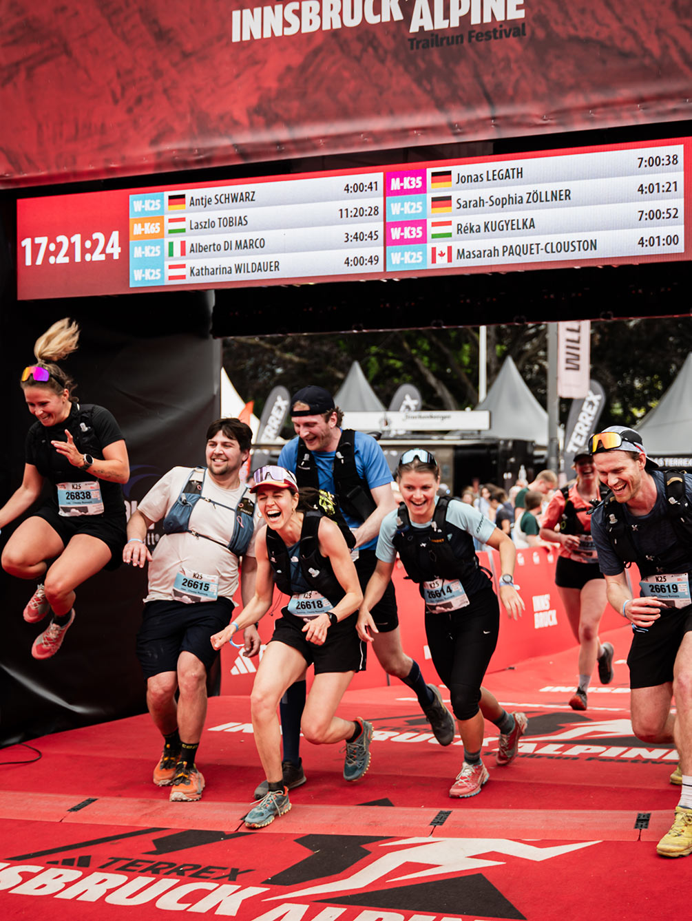 Eine jubelnde Gruppe von Trailrunnern läuft gemeinsam über die Ziellinie beim Innsbruck Alpine Trailrun Festival, während eine Läuferin vor Freude in die Luft springt.