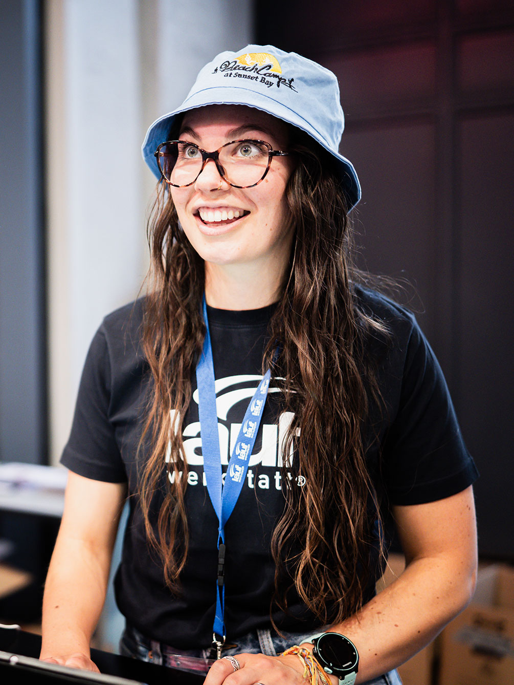 Eine lächelnde Mitarbeiterin des IATF-Teams mit Brille, blauem Bucket Hat und Laufwerkstatt-Shirt steht bereit für die Startnummernausgabe beim Innsbruck Alpine Trailrun Festival.