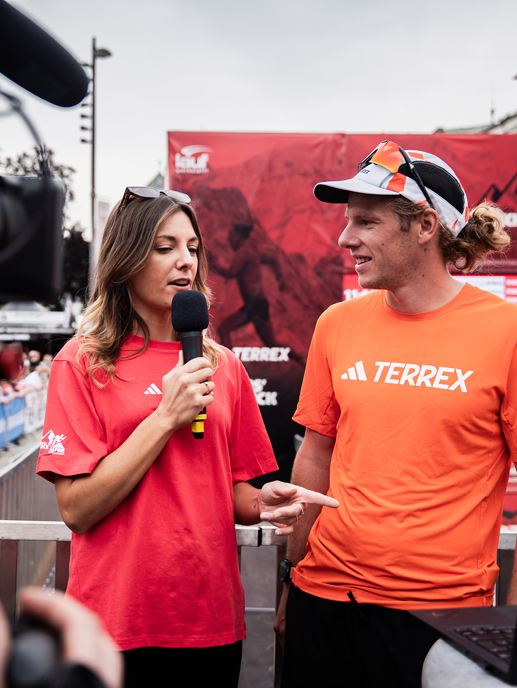Eine Moderatorin im roten Event-Shirt interviewt einen Trailrunner im orangefarbenen Terrex-Shirt vor dem Zielbogen beim Innsbruck Alpine Trailrun Festival.